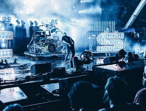A live concert scene features a vibrant stage with intense lighting and fog effects. Visible are large illuminated letters, a musician playing guitar, and a drum set. The audience is partially visible in the foreground, creating an energetic atmosphere.