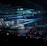 A large indoor arena filled with an audience watching a live performance. The stage features large screens displaying a performer and graphics, with bright spotlights and ambient lighting creating a dynamic atmosphere. The crowd is holding glow sticks, adding vibrant colors to the scene.