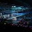 A large indoor arena filled with an audience watching a live performance. The stage features large screens displaying a performer and graphics, with bright spotlights and ambient lighting creating a dynamic atmosphere. The crowd is holding glow sticks, adding vibrant colors to the scene.