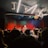 A live music performance in a dimly lit venue, with a vibrant curtain backdrop. A singer is performing on stage, accompanied by musicians. The audience, some recording with their phones, watches closely from the foreground. Stage lights create a dynamic and colorful atmosphere.