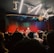 A live music performance in a dimly lit venue, with a vibrant curtain backdrop. A singer is performing on stage, accompanied by musicians. The audience, some recording with their phones, watches closely from the foreground. Stage lights create a dynamic and colorful atmosphere.