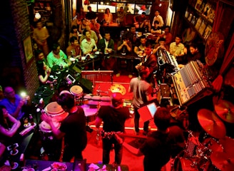 A lively music venue filled with people enjoying a live performance. The setting features a band with various musical instruments like drums, keyboards, and guitars on a stage with red lighting. The audience is seated closely around the stage, providing an intimate atmosphere. The room is dimly lit with warm, ambient lighting, enhancing the cozy and energetic vibe of the event.