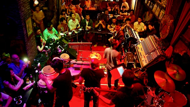 A lively music venue filled with people enjoying a live performance. The setting features a band with various musical instruments like drums, keyboards, and guitars on a stage with red lighting. The audience is seated closely around the stage, providing an intimate atmosphere. The room is dimly lit with warm, ambient lighting, enhancing the cozy and energetic vibe of the event.
