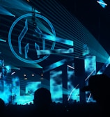 An indoor music event featuring a DJ booth illuminated with vibrant blue laser lights and large visual screens displaying abstract shapes. The crowd is silhouetted against the bright stage, adding to the energetic atmosphere.