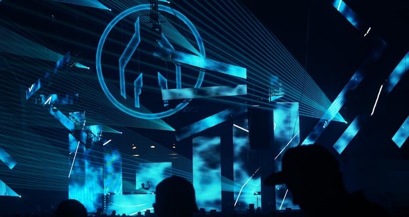 An indoor music event featuring a DJ booth illuminated with vibrant blue laser lights and large visual screens displaying abstract shapes. The crowd is silhouetted against the bright stage, adding to the energetic atmosphere.