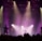 A live music performance with musicians silhouetted against a backdrop of bright stage lights. The scene is framed by beams of purple and yellow light, which enhance the dramatic mood of the concert. A crowd is visible in the foreground, adding to the lively atmosphere.