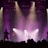 A live music performance with musicians silhouetted against a backdrop of bright stage lights. The scene is framed by beams of purple and yellow light, which enhance the dramatic mood of the concert. A crowd is visible in the foreground, adding to the lively atmosphere.