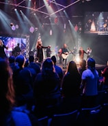 A live music performance takes place on a stage with a band and singers under bright, colorful stage lights. Audience members are standing in a dimly lit venue, watching and enjoying the performance. Decorations are visible, suggesting a festive or concert atmosphere, and a large screen displays visuals and lyrics.