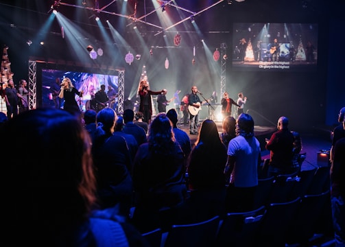 A live music performance takes place on a stage with a band and singers under bright, colorful stage lights. Audience members are standing in a dimly lit venue, watching and enjoying the performance. Decorations are visible, suggesting a festive or concert atmosphere, and a large screen displays visuals and lyrics.
