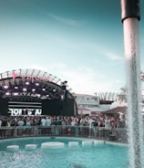 A large outdoor music event taking place in front of a stage with bright lights and a prominent sign above. The crowd is gathered around a swimming pool with water features and circular seats. People are enjoying the vibrant atmosphere, with modern architecture and palm trees in the background.