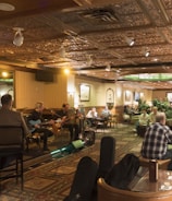 A warmly lit interior of a bar or restaurant featuring a live band performing on one side. The audience, seated at tables and bar stools, enjoys the music. The space is decorated with intricate ceiling designs and a mounted animal head on the wall. The atmosphere is cozy and lively with social interactions throughout.