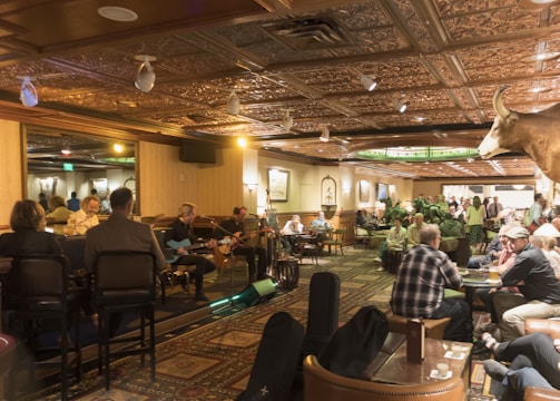 A warmly lit interior of a bar or restaurant featuring a live band performing on one side. The audience, seated at tables and bar stools, enjoys the music. The space is decorated with intricate ceiling designs and a mounted animal head on the wall. The atmosphere is cozy and lively with social interactions throughout.