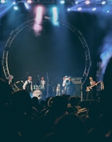 A live music performance on stage with several musicians playing instruments including drums and guitar. The stage is illuminated with colorful lights and smoke effects, creating a lively and dynamic atmosphere. The audience is visible in silhouette, indicating an engaged crowd.