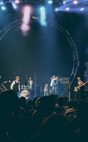 A live music performance on stage with several musicians playing instruments including drums and guitar. The stage is illuminated with colorful lights and smoke effects, creating a lively and dynamic atmosphere. The audience is visible in silhouette, indicating an engaged crowd.