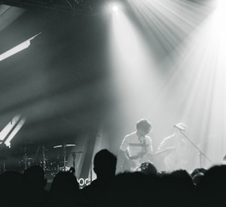 A live music performance with musicians playing on stage, surrounded by dramatic lighting and a crowd in the foreground. The atmosphere is dynamic, with beams of light cutting through hazy air, creating an intense and immersive experience.