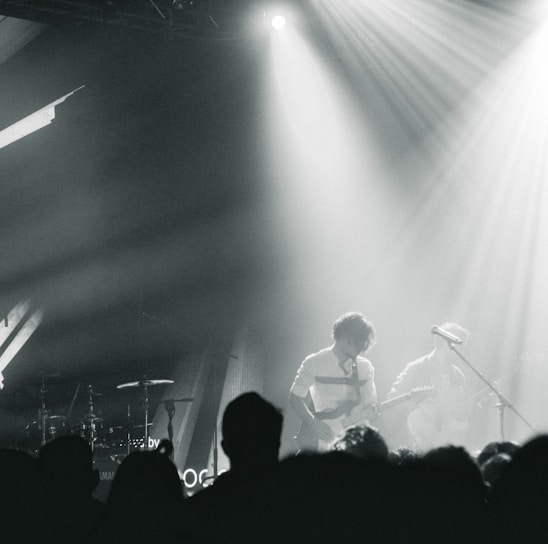 A live music performance with musicians playing on stage, surrounded by dramatic lighting and a crowd in the foreground. The atmosphere is dynamic, with beams of light cutting through hazy air, creating an intense and immersive experience.