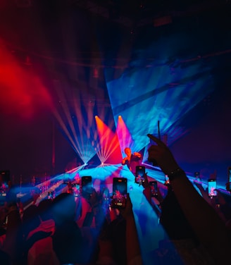 A live concert event with vibrant lighting effects. The stage is bathed in a mix of blue and red lights, creating a striking visual. The crowd in the foreground holds up smartphones, capturing the performance. The performer stands at the center of the stage, illuminated by spotlights.