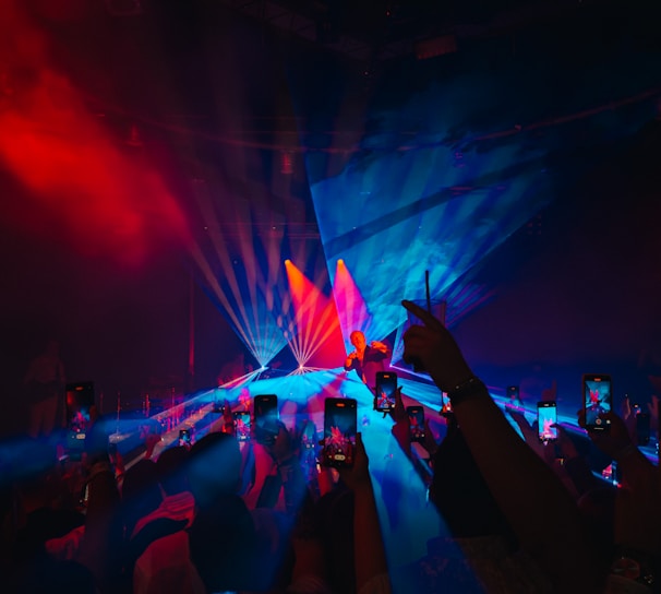 A live concert event with vibrant lighting effects. The stage is bathed in a mix of blue and red lights, creating a striking visual. The crowd in the foreground holds up smartphones, capturing the performance. The performer stands at the center of the stage, illuminated by spotlights.