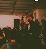 A live music performance is taking place in a compact indoor venue with a crowd of people gathered around. The lead singer is passionately holding a microphone and singing, surrounded by audience members who appear engaged and energetic. The setting is dimly lit, creating an intimate ambiance.