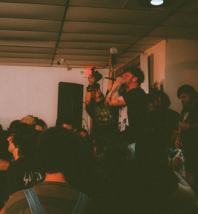 A live music performance is taking place in a compact indoor venue with a crowd of people gathered around. The lead singer is passionately holding a microphone and singing, surrounded by audience members who appear engaged and energetic. The setting is dimly lit, creating an intimate ambiance.