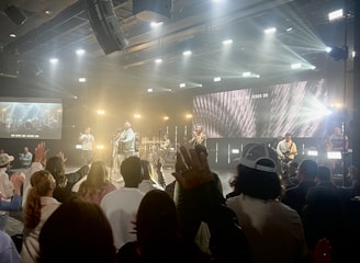 A live music event with a band performing on stage. Bright lights and multimedia screens illuminate the space, creating a lively atmosphere. The audience is standing with several people raising their hands, suggesting engagement and enthusiasm.