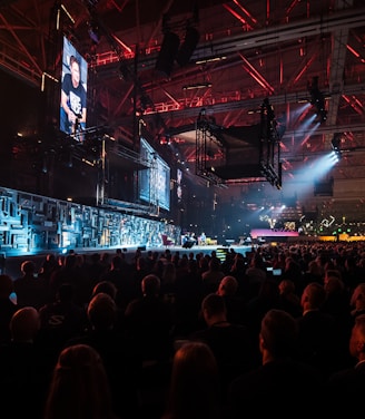 A large indoor event with a stage featuring bright screens and spotlights, attended by a substantial crowd. The venue has a high ceiling with industrial beams and red lighting accents. The atmosphere resembles a concert or a conference with a focus on visual effects.