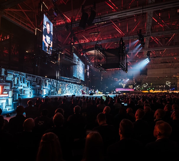 A large indoor event with a stage featuring bright screens and spotlights, attended by a substantial crowd. The venue has a high ceiling with industrial beams and red lighting accents. The atmosphere resembles a concert or a conference with a focus on visual effects.