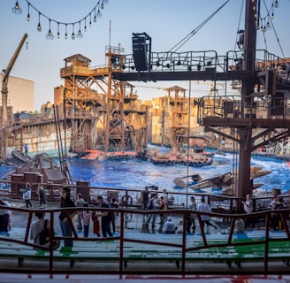 An outdoor amphitheater with a large pool of water is set up for a live show. The stage is designed to resemble an industrial or post-apocalyptic world with elaborate metal structures, scaffolding, and decorative elements. The audience is composed of people of various ages, casually dressed, and seated or standing, eagerly anticipating the performance. Overhead, string lights add to the setting's ambiance.
