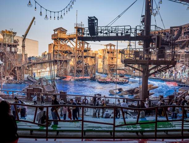An outdoor amphitheater with a large pool of water is set up for a live show. The stage is designed to resemble an industrial or post-apocalyptic world with elaborate metal structures, scaffolding, and decorative elements. The audience is composed of people of various ages, casually dressed, and seated or standing, eagerly anticipating the performance. Overhead, string lights add to the setting's ambiance.