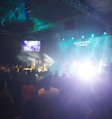 A live performance is taking place in a dimly lit indoor space. The room is filled with people watching the stage, where several performers are illuminated by bright lights against a blue and green backdrop. There are screens on the sides showing the stage activity. Smoke or fog effects enhance the dramatic lighting on stage.