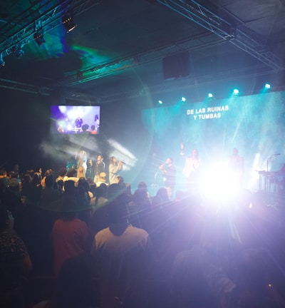 A live performance is taking place in a dimly lit indoor space. The room is filled with people watching the stage, where several performers are illuminated by bright lights against a blue and green backdrop. There are screens on the sides showing the stage activity. Smoke or fog effects enhance the dramatic lighting on stage.