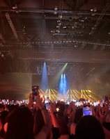 A dimly lit indoor concert venue filled with an audience holding up lit devices and lantern-like objects, capturing a performance on stage. Bright spotlights and blue and yellow beams illuminate the stage area, creating a lively atmosphere.