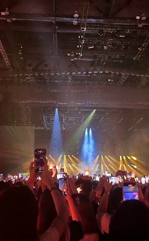 A dimly lit indoor concert venue filled with an audience holding up lit devices and lantern-like objects, capturing a performance on stage. Bright spotlights and blue and yellow beams illuminate the stage area, creating a lively atmosphere.