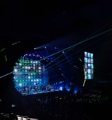 A concert stage is illuminated with vibrant lights and screens displaying various images. The atmosphere is dynamic, with beams of light cutting across the venue filled with a large audience. The stage is equipped with multiple screens and a circular arrangement of lights, creating an immersive visual experience.