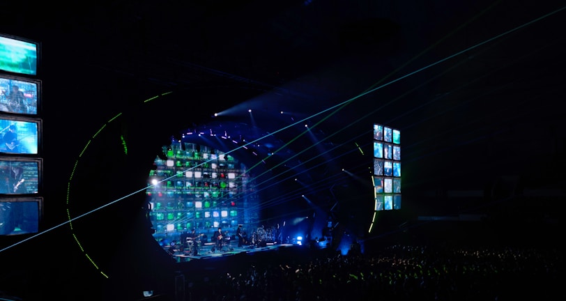A concert stage is illuminated with vibrant lights and screens displaying various images. The atmosphere is dynamic, with beams of light cutting across the venue filled with a large audience. The stage is equipped with multiple screens and a circular arrangement of lights, creating an immersive visual experience.