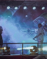 A dimly lit stage features performers surrounded by colorful fog, with bright stage lights overhead. The backdrop displays large images of beverage bottles, creating a lively and energetic atmosphere.