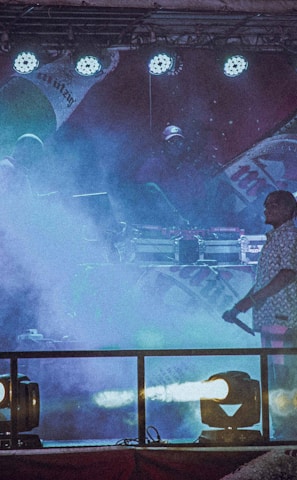 A dimly lit stage features performers surrounded by colorful fog, with bright stage lights overhead. The backdrop displays large images of beverage bottles, creating a lively and energetic atmosphere.